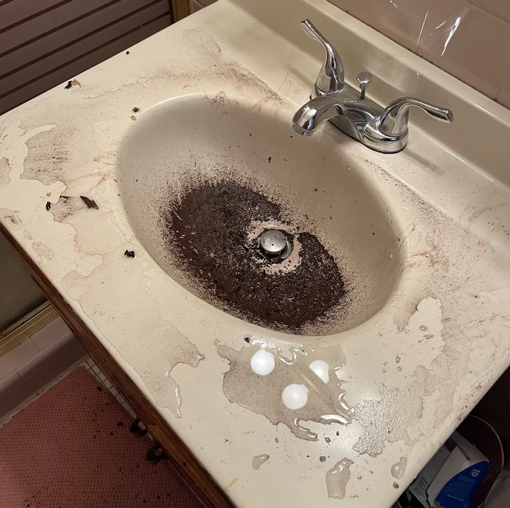 Clogged bathroom sink with dark drain debris and standing residue around the basin, showing a messy backup related to how to unclog a bathroom sink.